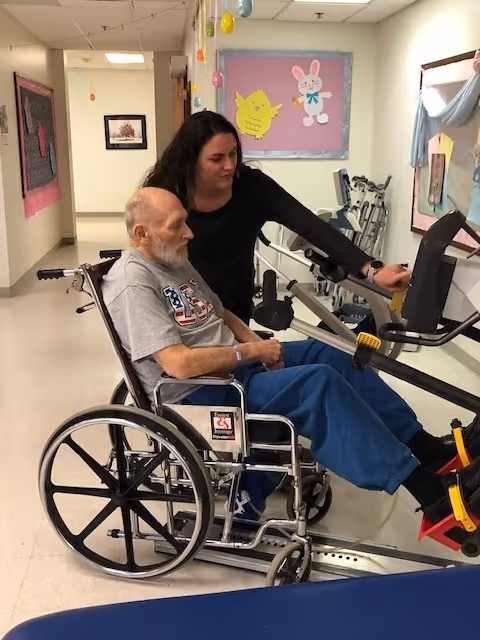 An elderly man in a wheelchair is using a piece of exercise equipment with the assistance of a woman standing beside him in a hallway decorated with colorful paper cutouts of a chick and a bunny on the walls.