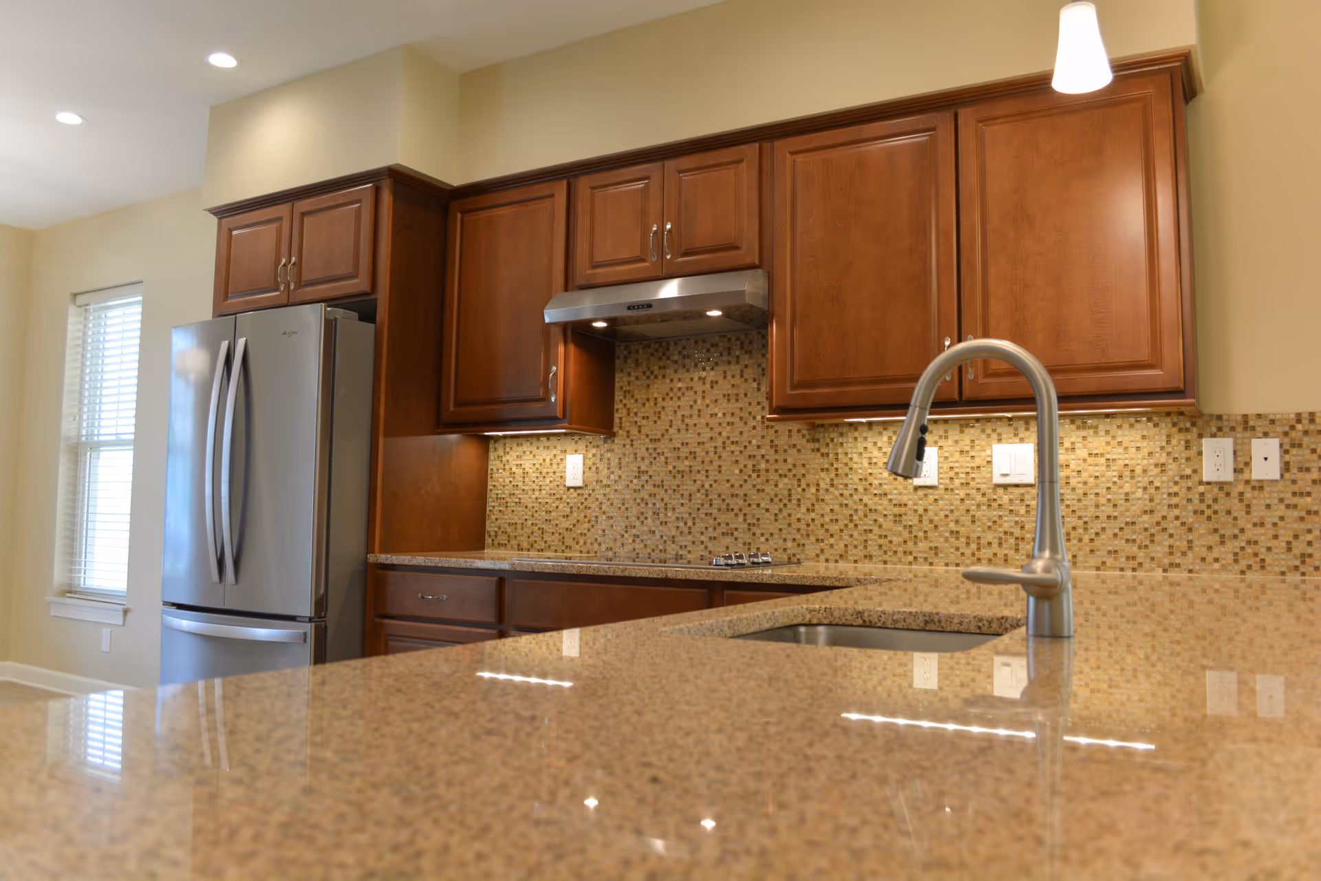 Modern kitchen with brown wooden cabinets, a stainless steel refrigerator, a mosaic tile backsplash, and a granite countertop with a built-in sink and a curved faucet.