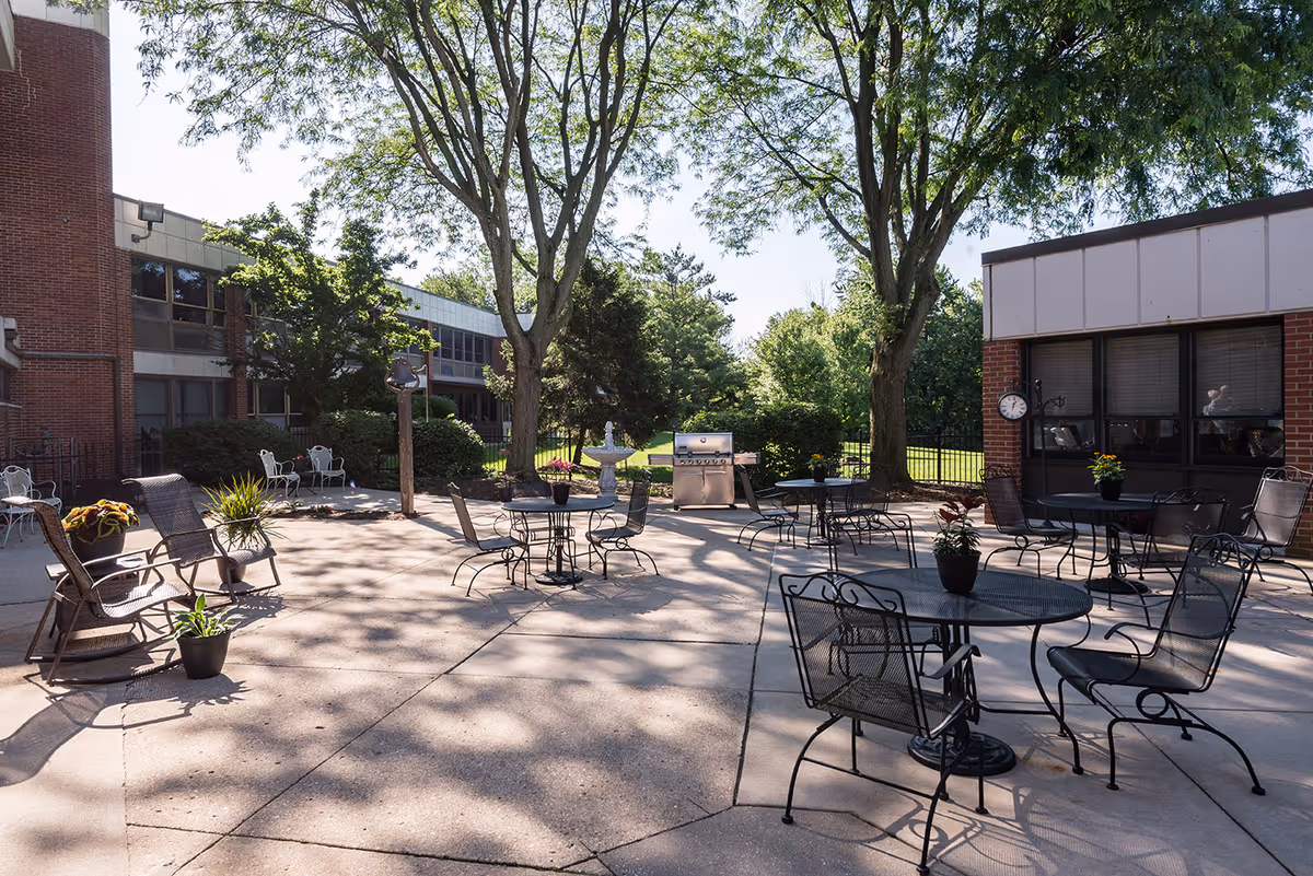Outdoor patio area at Altenheim Family-first Senior Living with several metal tables and chairs, potted plants on the tables, a barbecue grill, and large trees providing shade. The patio is surrounded by brick buildings and greenery.