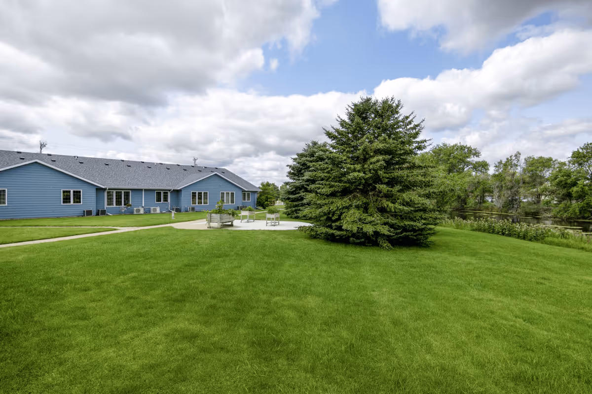 A large green lawn with a cluster of evergreen trees in the center, a concrete patio area with benches, and a blue single-story building in the background under a partly cloudy sky.