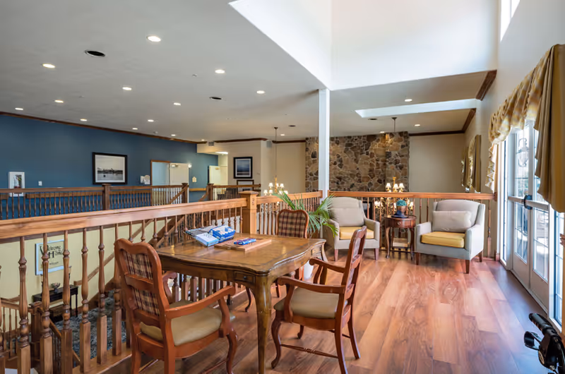 A bright and spacious interior common area with wooden flooring, a wooden table with four chairs, two cushioned armchairs with yellow seat cushions, a small round side table with a plant, and large windows with yellow curtains allowing natural light to fill the space. The area features a stone accent wall and wooden railing overlooking a lower level.