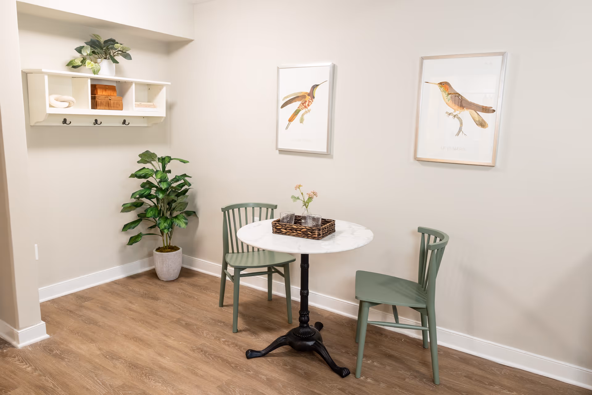 A small dining area with a round white marble table and two green wooden chairs. On the table is a woven tray holding two glasses and a small vase with flowers. The walls are light beige and decorated with two framed bird illustrations. A white wall-mounted shelf with hooks holds decorative items, and a potted green plant is placed in the corner on a wooden floor.