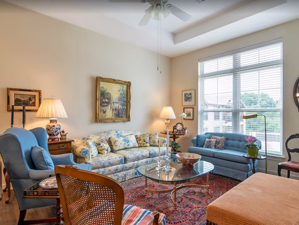 Bright, traditionally furnished living room with a floral sofa, blue armchairs, glass coffee table, patterned rug and a large window.