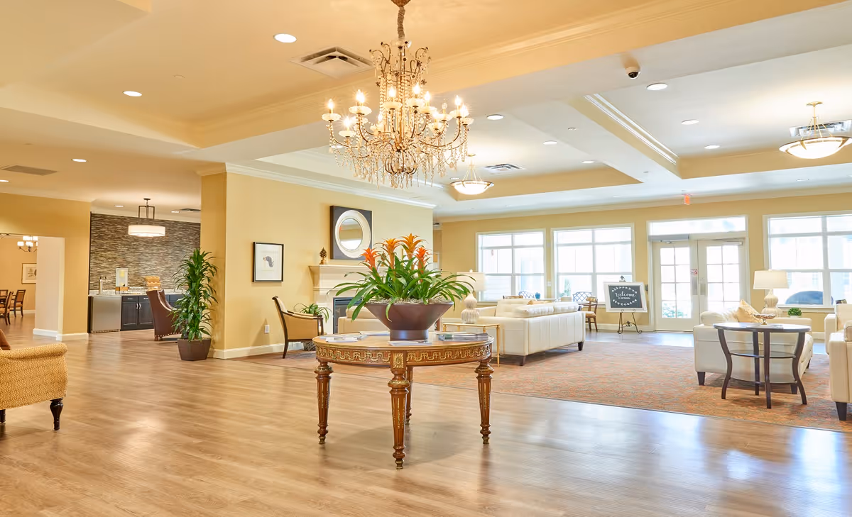 A spacious and well-lit senior living facility common area with wooden flooring, a chandelier, a round table with a flower arrangement in the center, comfortable white sofas, armchairs, large windows, and a welcome sign near the entrance door.