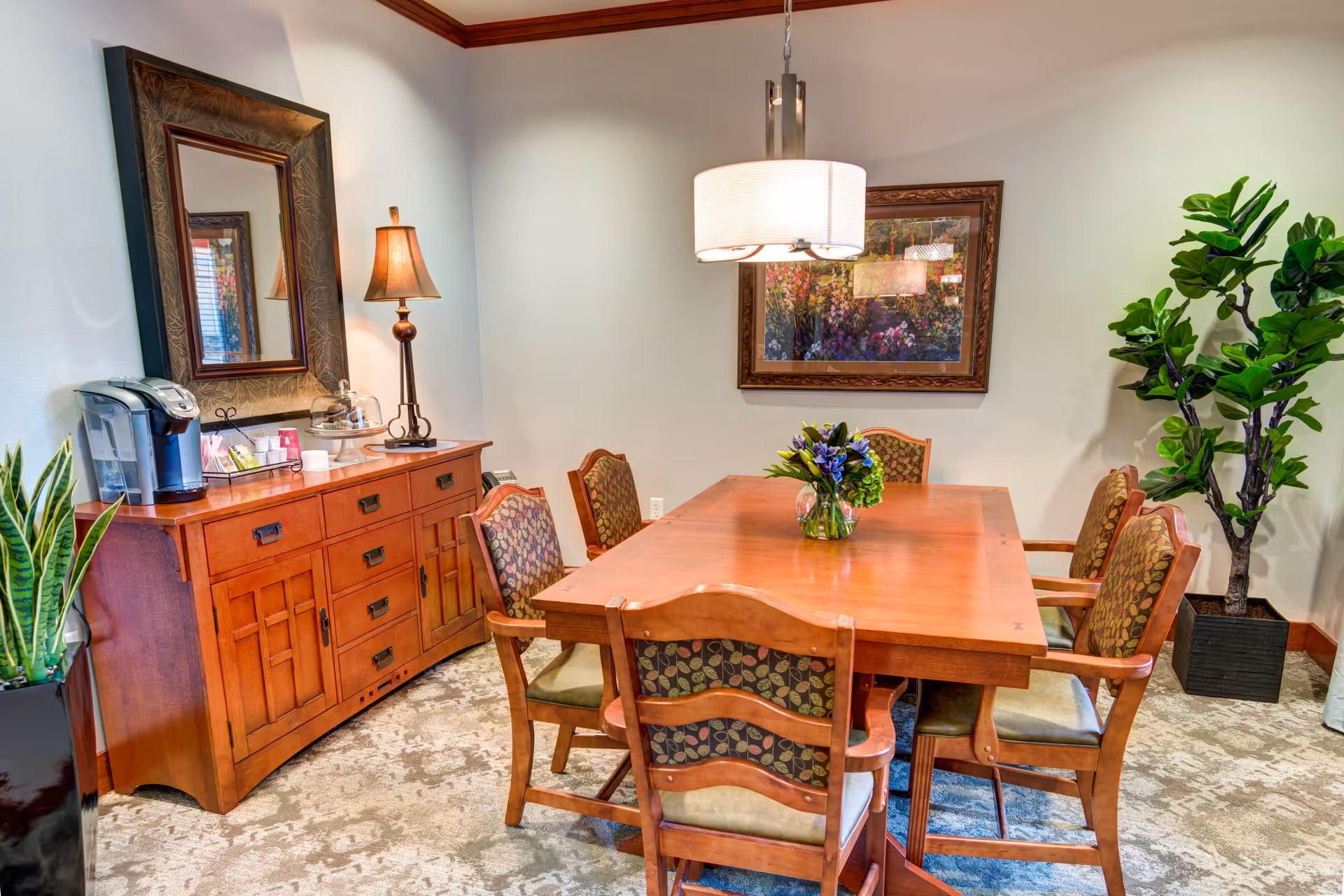 A dining room with a wooden table surrounded by six cushioned chairs with patterned backs. On the table is a small vase with flowers. Against the wall is a wooden sideboard with a lamp, a coffee maker, and various items. Above the sideboard is a large framed mirror, and on the wall opposite the sideboard is a framed painting. A large potted plant is in the corner, and a hanging light fixture is above the table.