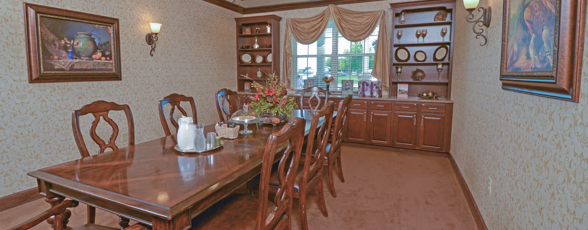 A formal dining room with a long wooden dining table surrounded by eight matching wooden chairs. The table has a centerpiece with flowers, a glass cake stand, a pitcher, and a water carafe. The room features patterned wallpaper, two framed paintings on the walls, wall sconces with lights, and built-in wooden cabinets and shelves displaying decorative plates and glassware. A window with draped curtains lets in natural light.