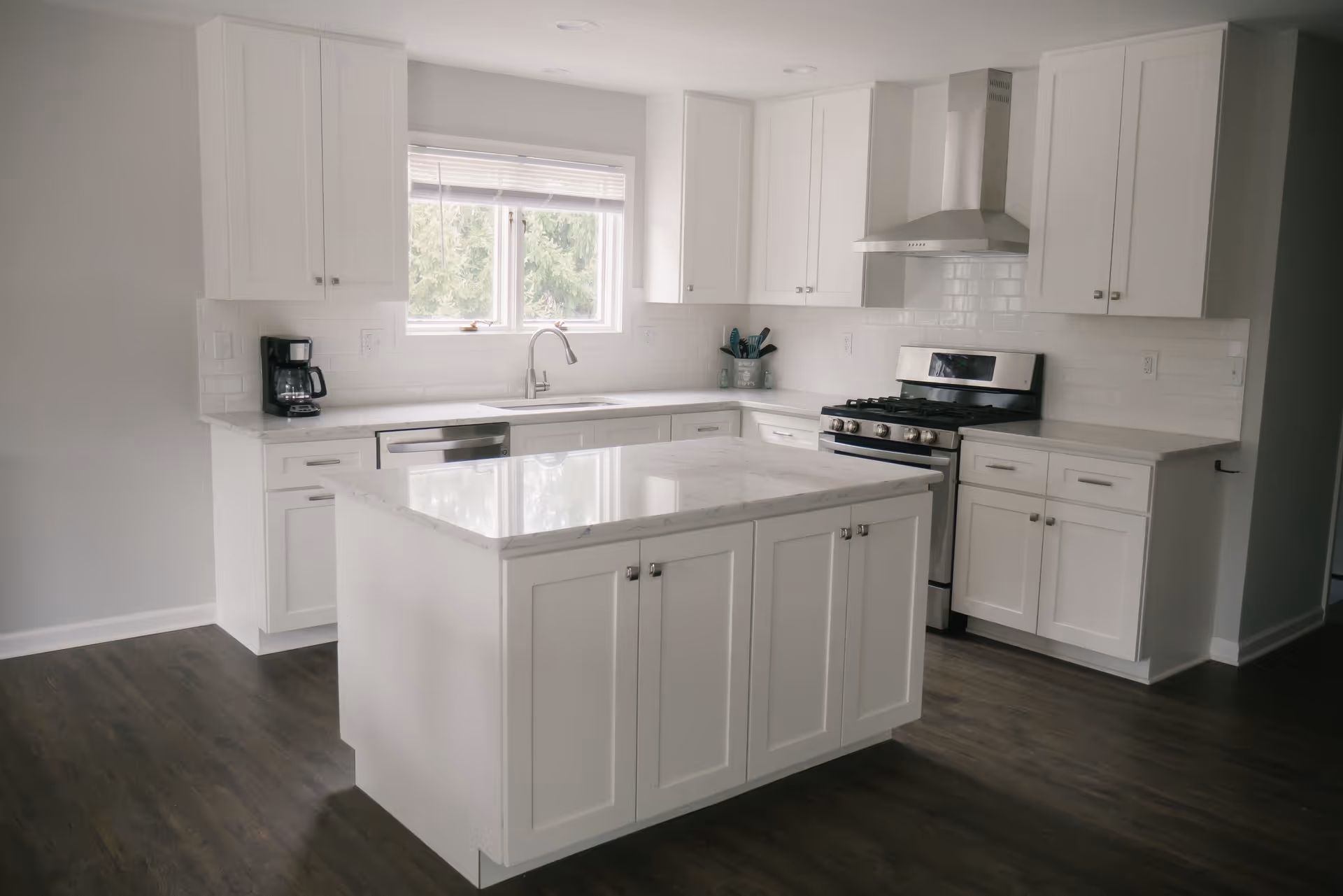A modern kitchen with white cabinets and drawers, a central island with a marble countertop, a stainless steel stove with a hood, a dishwasher, a coffee maker, and a window above the sink showing greenery outside. The floor is dark wood.