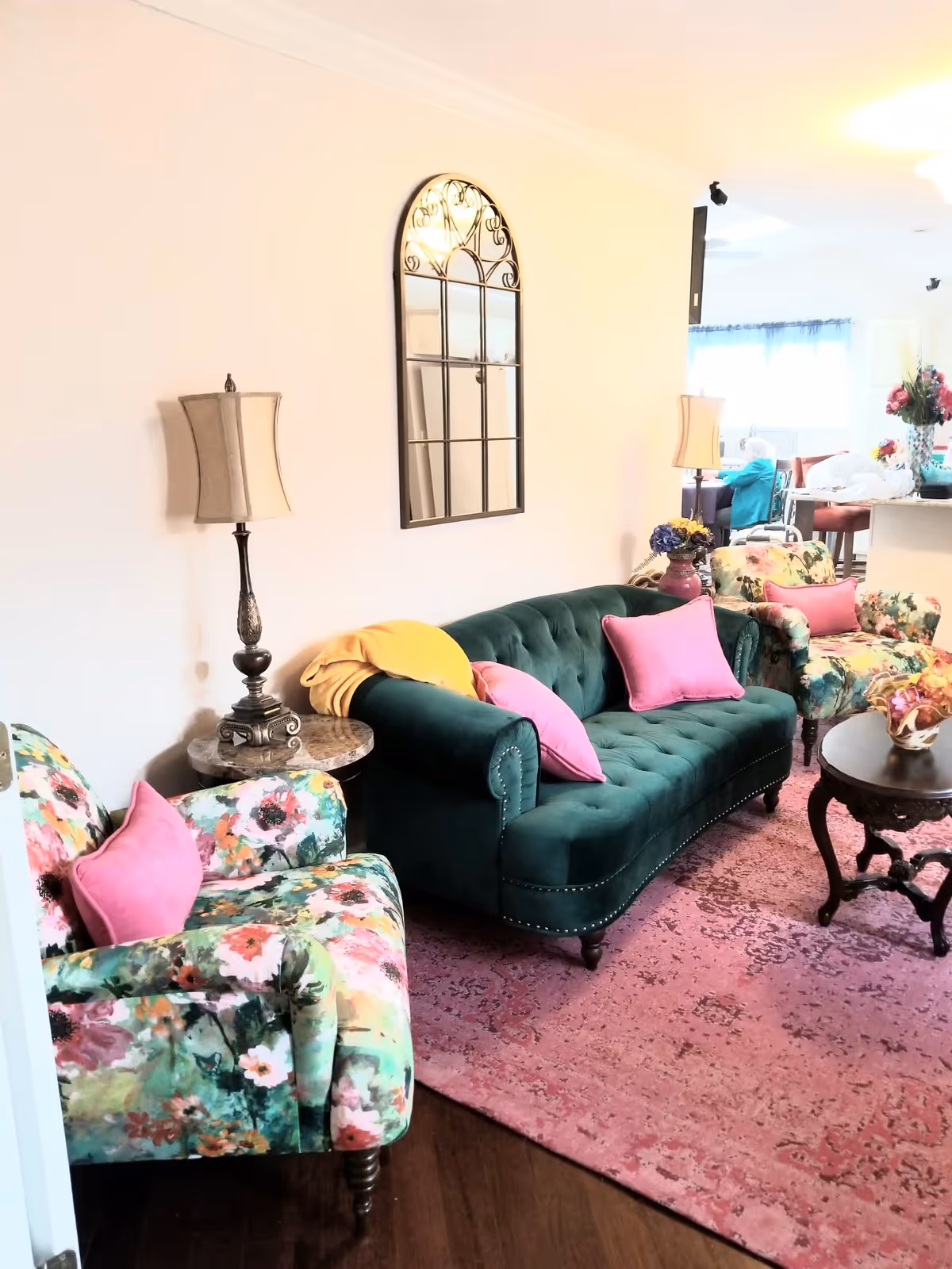 Bright living room with a green velvet sofa, floral armchairs, pink pillows, lamps, a wall mirror, and a pink area rug.