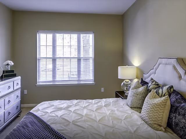 Bright bedroom with a bed dressed in decorative pillows and a lamp-lit nightstand beside a window with blinds.