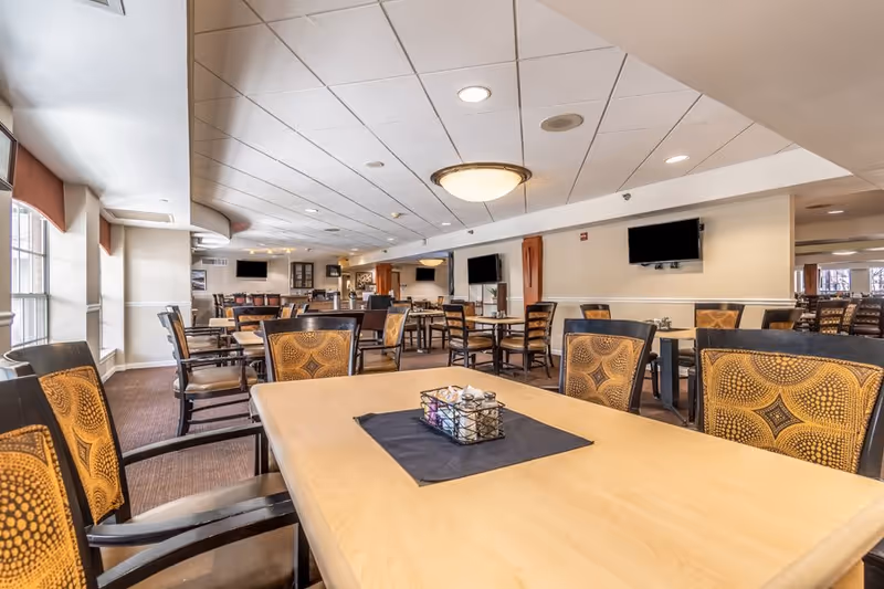A spacious dining room with multiple wooden tables and chairs featuring patterned upholstery. The room has large windows on one side allowing natural light to enter, ceiling lights, and several wall-mounted flat-screen TVs. The overall decor is warm and inviting.