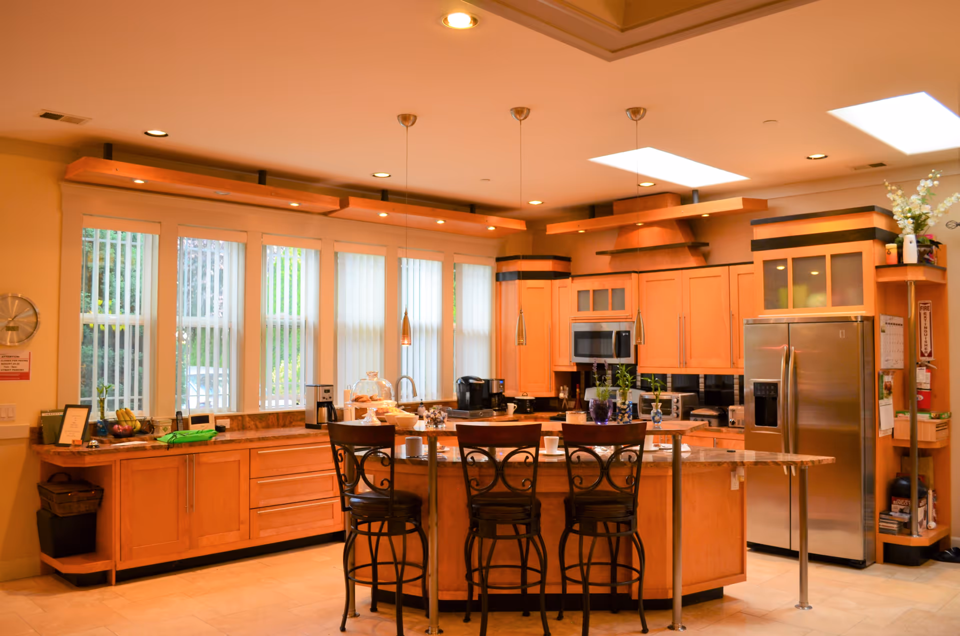 A spacious kitchen with wooden cabinets, a large island with three high chairs, pendant lights hanging above the island, stainless steel refrigerator, microwave, coffee maker, and large windows with vertical blinds allowing natural light to enter.