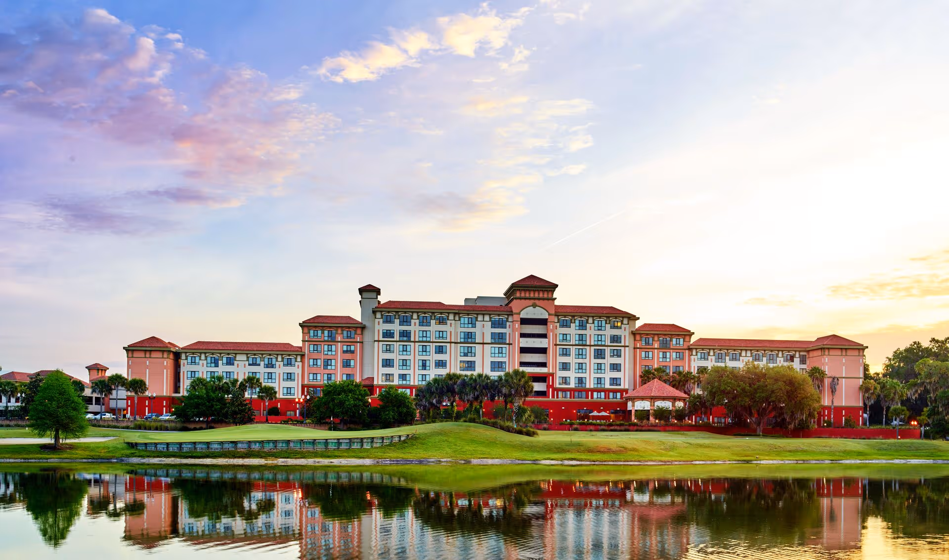 Wide exterior view of a large multi-story senior living facility named Freedom Pointe at The Villages, with a red and beige facade, surrounded by green lawns, trees, and a pond reflecting the building under a partly cloudy sky at sunset.