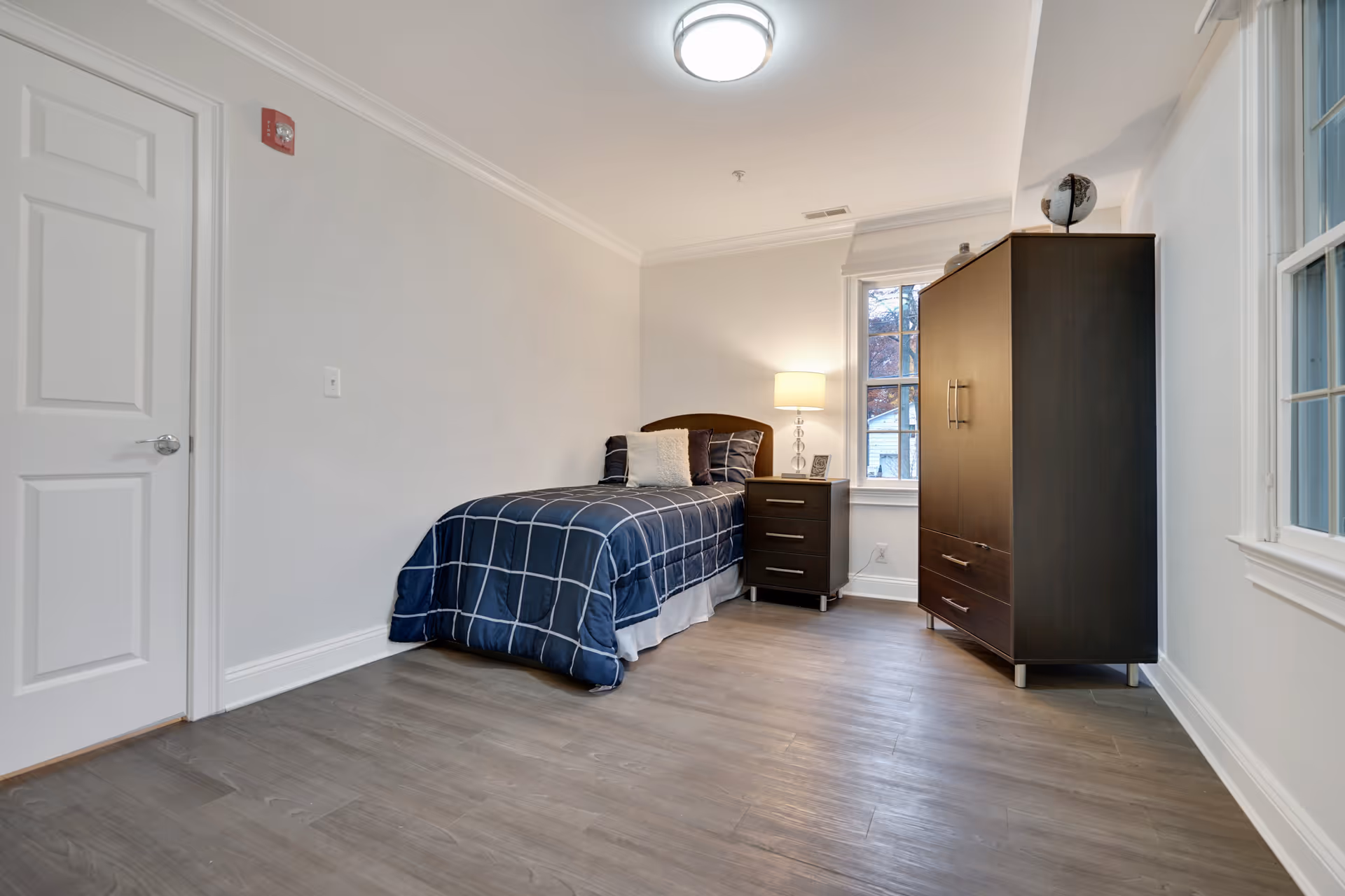 A simple bedroom with a single bed covered in a blue checkered comforter, a nightstand with a lamp and picture frame, a dark wooden wardrobe, two windows, and light-colored walls and flooring.
