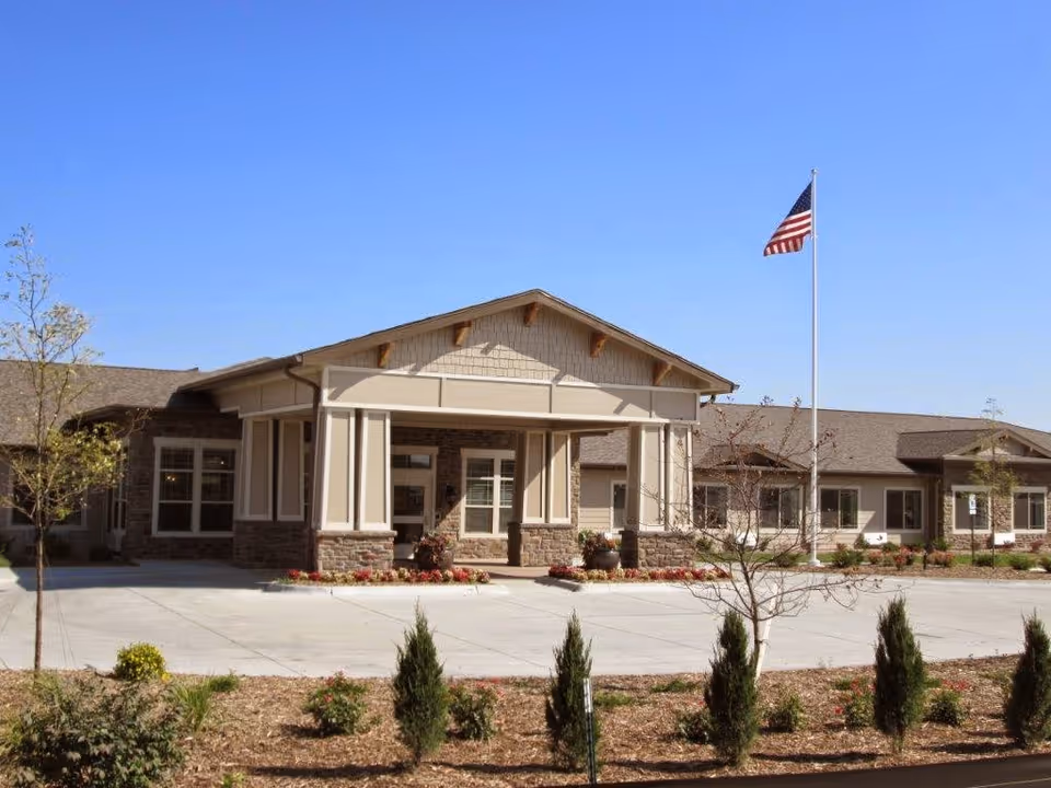 Exterior view of Cedar Creek of High Plains senior living facility showing a single-story building with a covered entrance, stone and beige siding, an American flag on a flagpole, and landscaped grounds with small trees and shrubs under a clear blue sky.