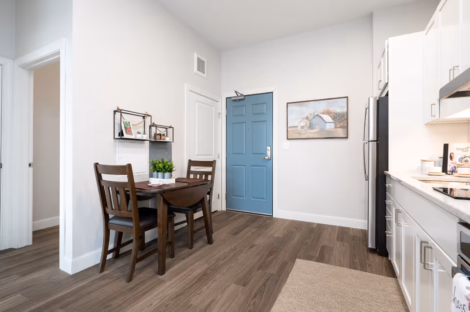 A small dining area with a dark wooden table and two chairs set against a white wall. The table has a small green plant and a cup on it. To the right is a kitchen area with white cabinets, a countertop, and a stainless steel refrigerator. A blue door and a framed painting of a house hang on the wall in the background. The floor is wood with a light-colored rug partially visible.