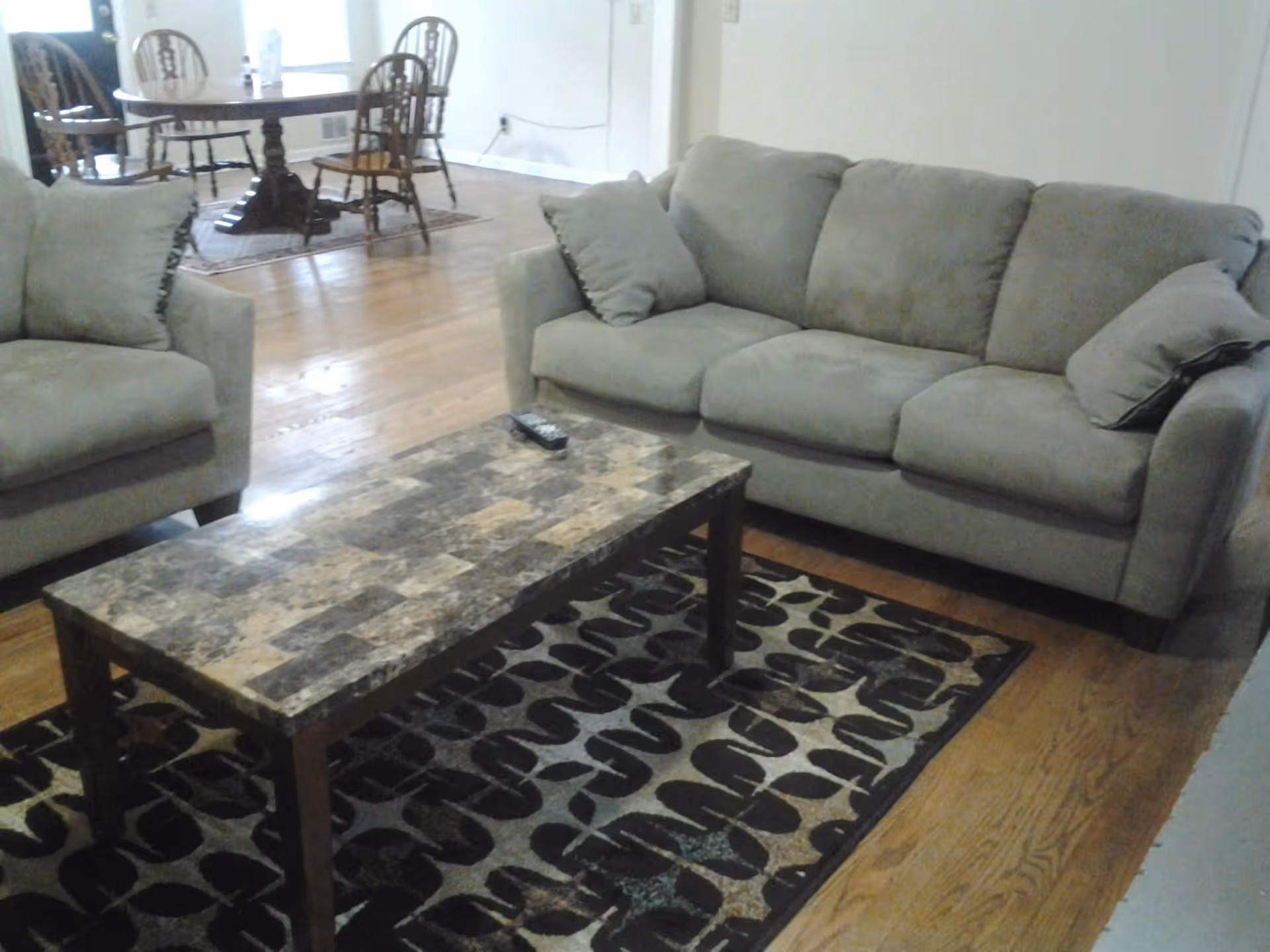 Living room with a gray sofa and armchair, a patterned coffee table and rug, and a dining table visible in the background.