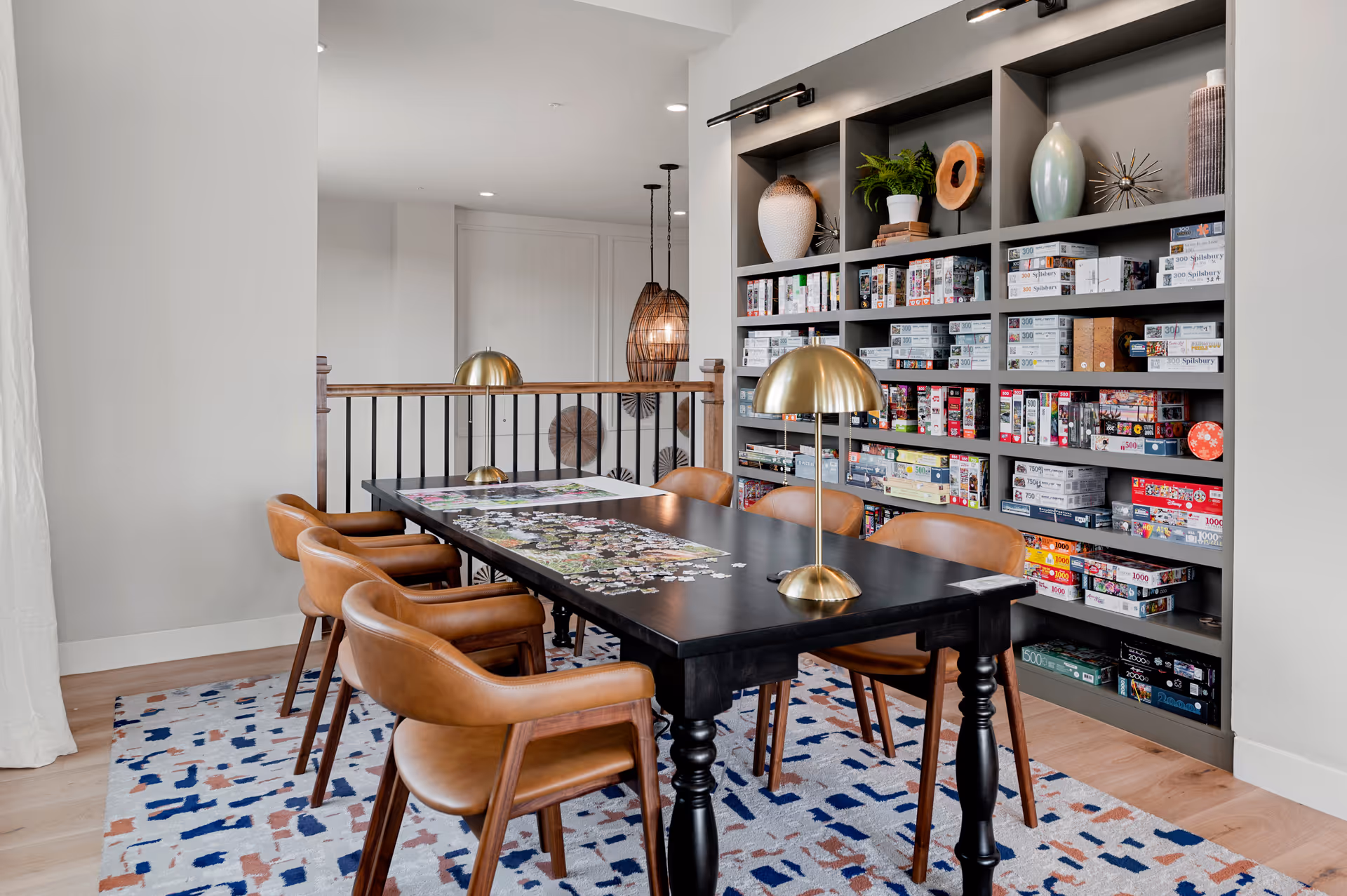 Modern activity room with a long black table surrounded by tan leather chairs, brass lamps, and shelving filled with board games and puzzles.