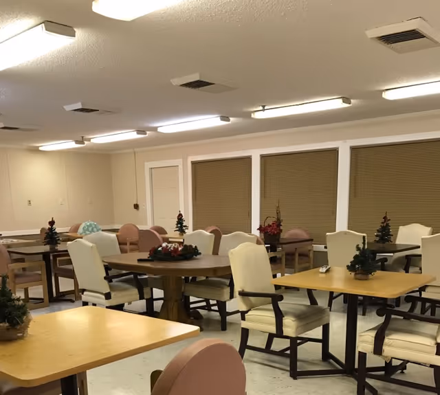 Communal dining room with round and square tables, upholstered chairs, and small holiday centerpieces under fluorescent lights.
