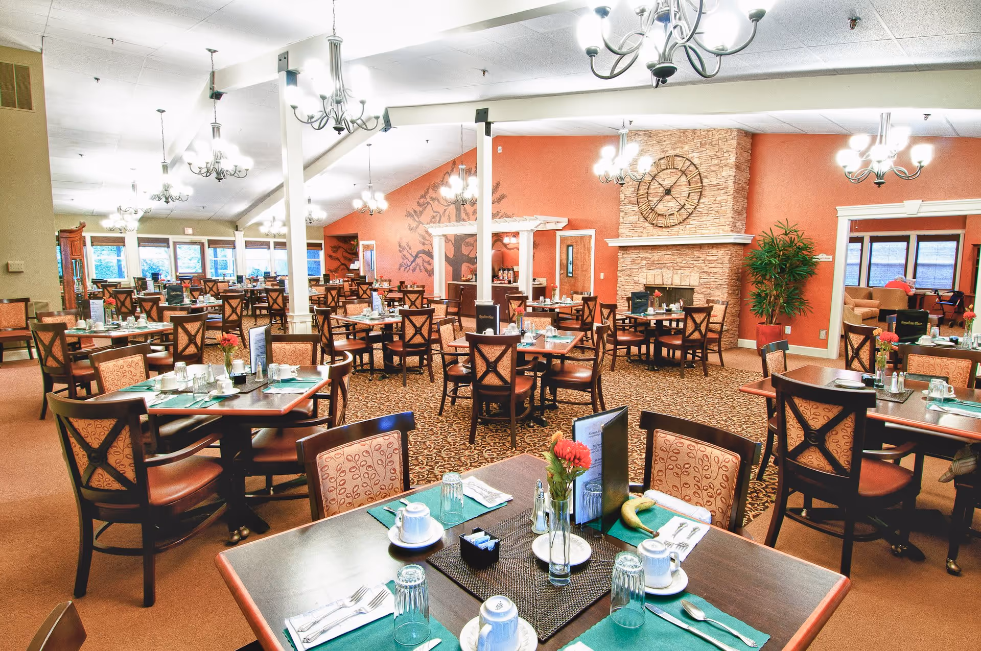 A spacious dining room with multiple tables and chairs arranged neatly. Each table is set with placemats, cups, glasses, utensils, and small flower vases. The room features warm lighting from chandeliers, a large clock mounted on a stone fireplace, and a mural on one wall. Large windows allow natural light to enter, and there is a cozy seating area visible in the background.