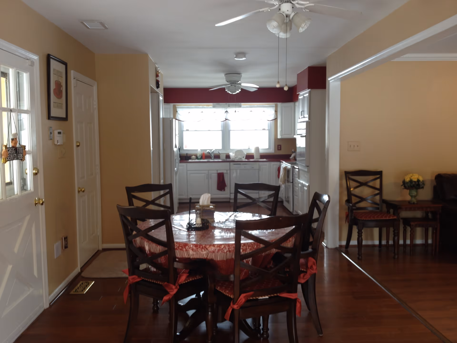 Interior view of a dining area with a round table covered with a red tablecloth and six wooden chairs with red cushions. The dining area opens into a kitchen with white cabinets, a refrigerator, a dishwasher, and a stove. The walls are painted beige with a red accent wall in the kitchen. There are ceiling fans with lights in both the dining and kitchen areas. To the right, there is a small sitting area with two wooden chairs and a side table with a flower vase.