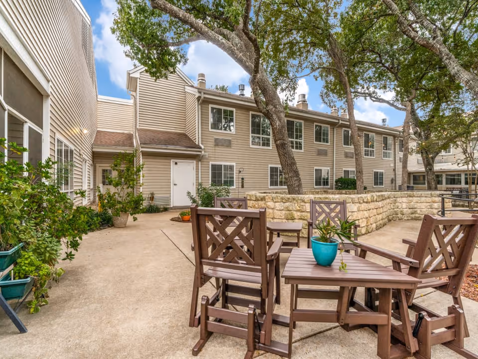 Outdoor patio area at Juniper Village at Spicewood Summit featuring wooden chairs and tables with a potted plant on one table, surrounded by trees and a stone retaining wall, adjacent to a beige two-story building with multiple windows.