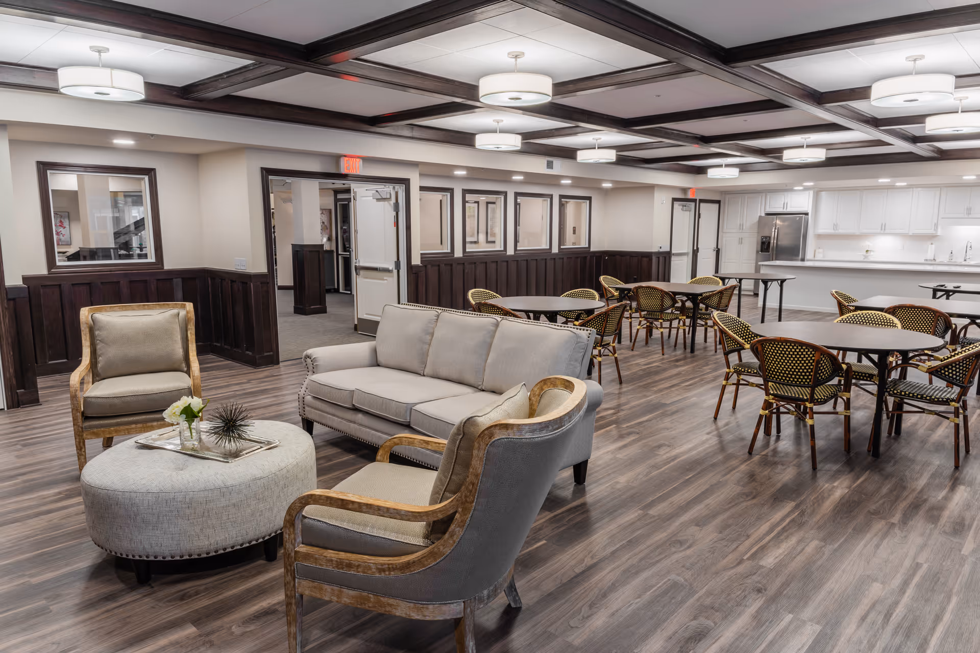 A spacious common area featuring a seating arrangement with a beige sofa, two armchairs, and a round ottoman with a decorative tray. In the background, there are several round tables with chairs arranged around them, and a kitchen area with white cabinetry and stainless steel appliances. The room has wood flooring, white ceiling lights, and dark wood trim accents.