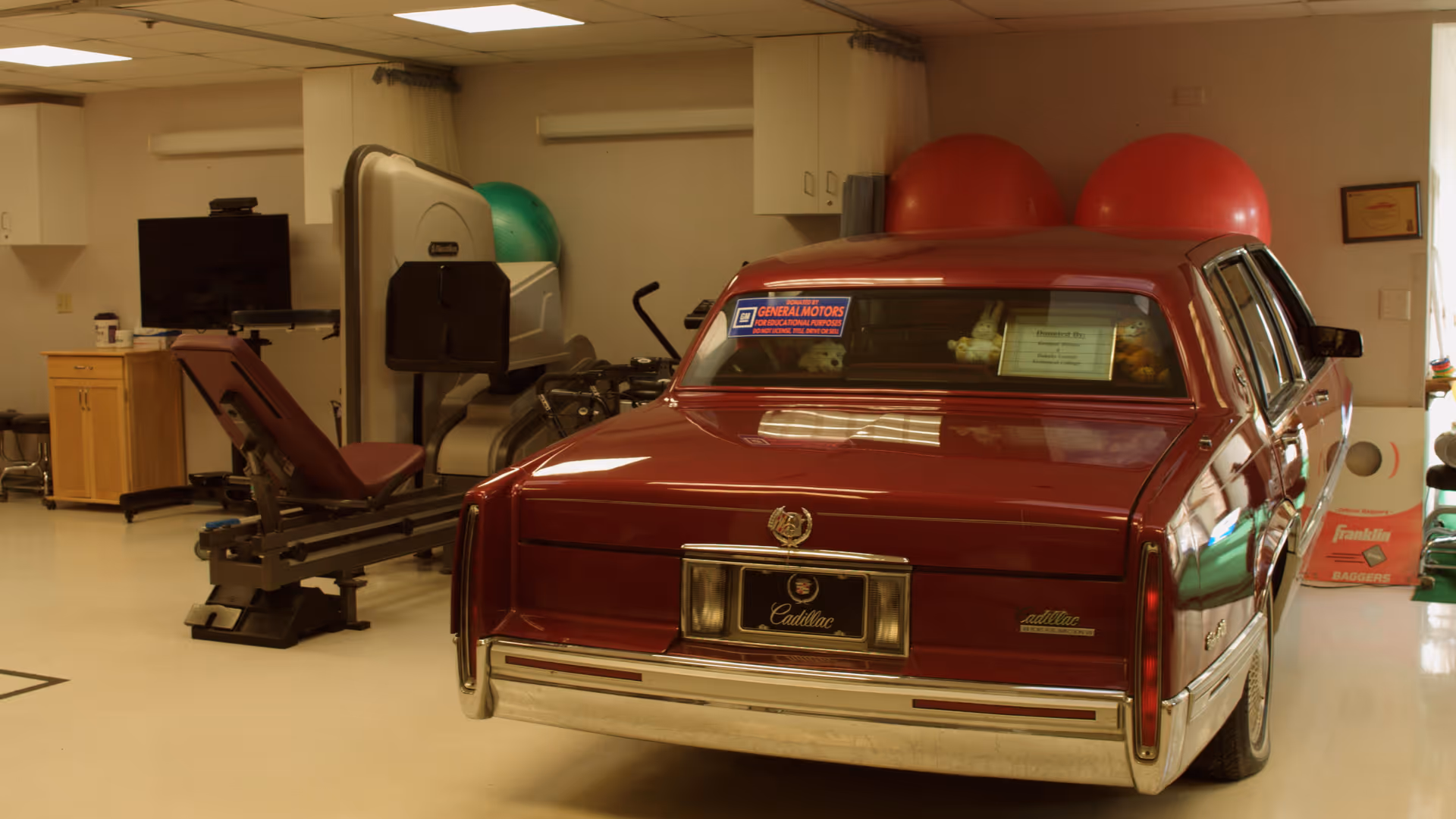 Interior room with exercise equipment including a leg press machine and large exercise balls, alongside a vintage red Cadillac car parked inside the room.