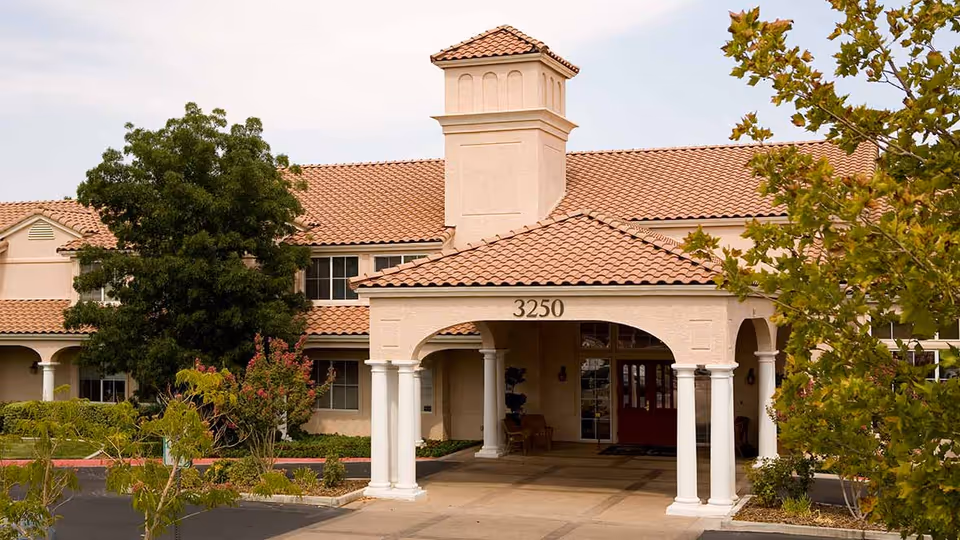 Exterior view of a senior living facility building with beige stucco walls and a red tiled roof. The entrance features a covered portico supported by white columns, with the number 3250 displayed above. There are trees and shrubs around the building and a paved driveway in front.