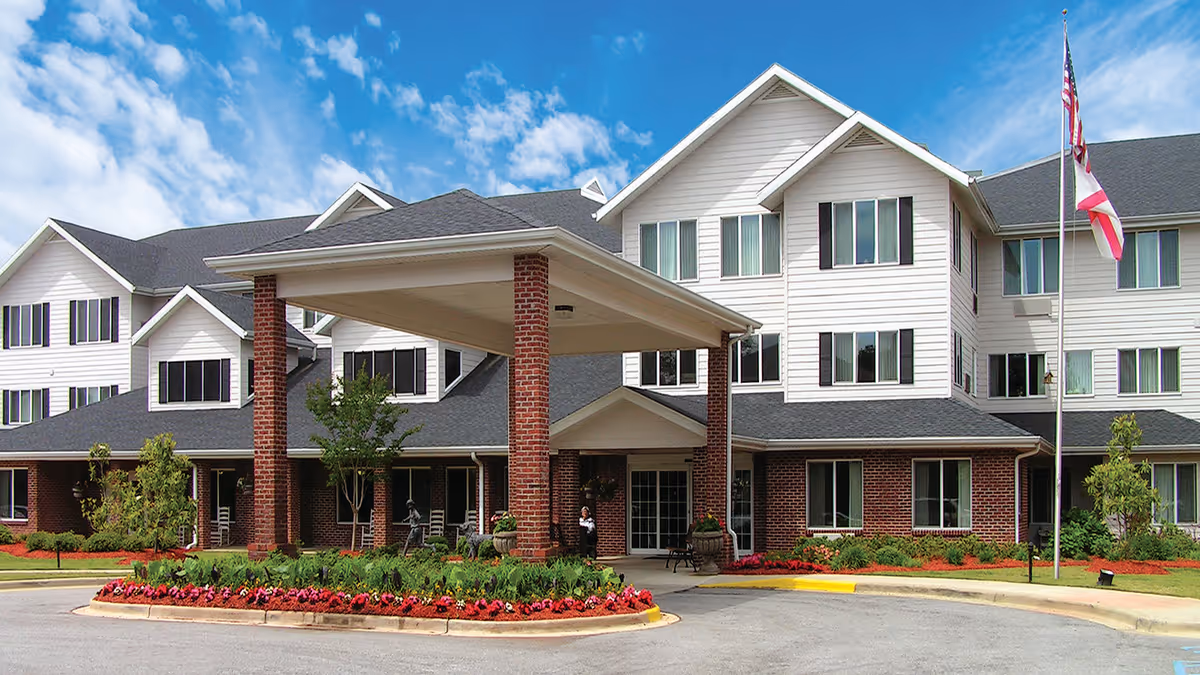 Front exterior view of a multi-story senior living facility building with white siding and brick accents, a covered entrance supported by brick columns, landscaped flower beds, and an American flag flying on a flagpole under a partly cloudy blue sky.