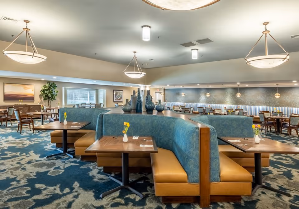 A spacious dining room with multiple wooden tables and cushioned booth seating upholstered in blue and tan. Each table has a small white vase with a yellow flower. The room features modern hanging light fixtures, a patterned carpet, and artwork on the walls. Large windows allow natural light to enter the space.