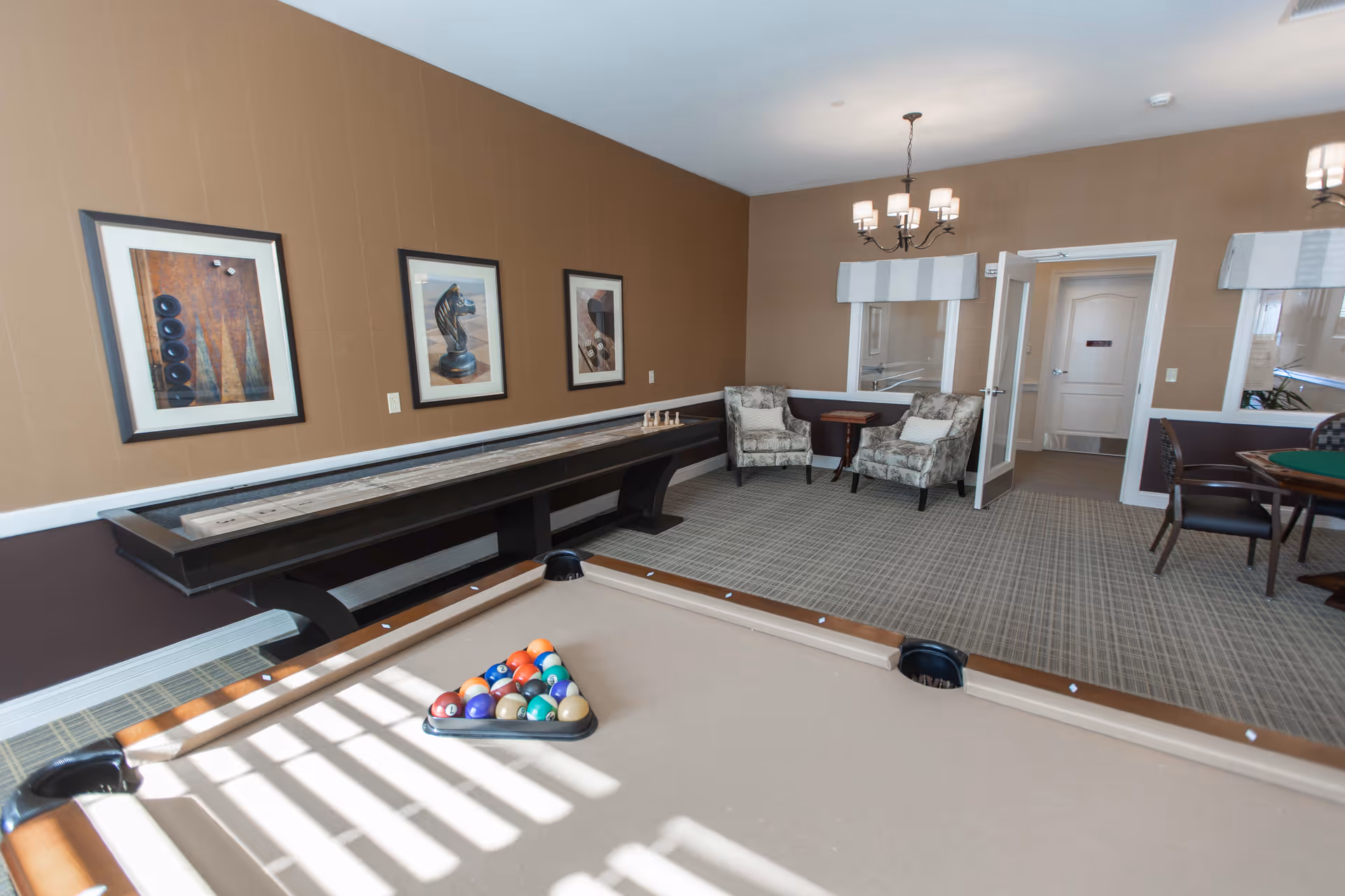 A recreational room with a pool table in the foreground, a shuffleboard table along the left wall, two patterned armchairs with a small table between them, and a card table with chairs on the right. The walls are painted brown with framed artwork, and the room is well-lit with natural light and ceiling chandeliers.