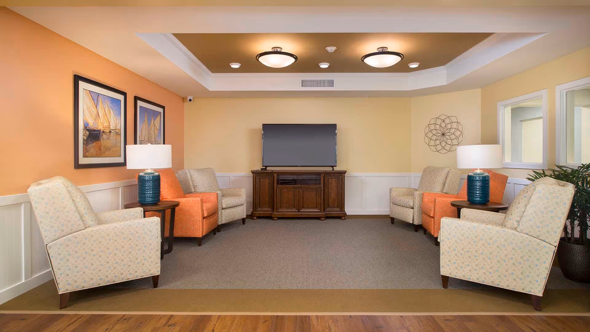 A furnished lounge with armchairs arranged around a TV on a wooden console, side tables with lamps, and framed artwork on the walls.