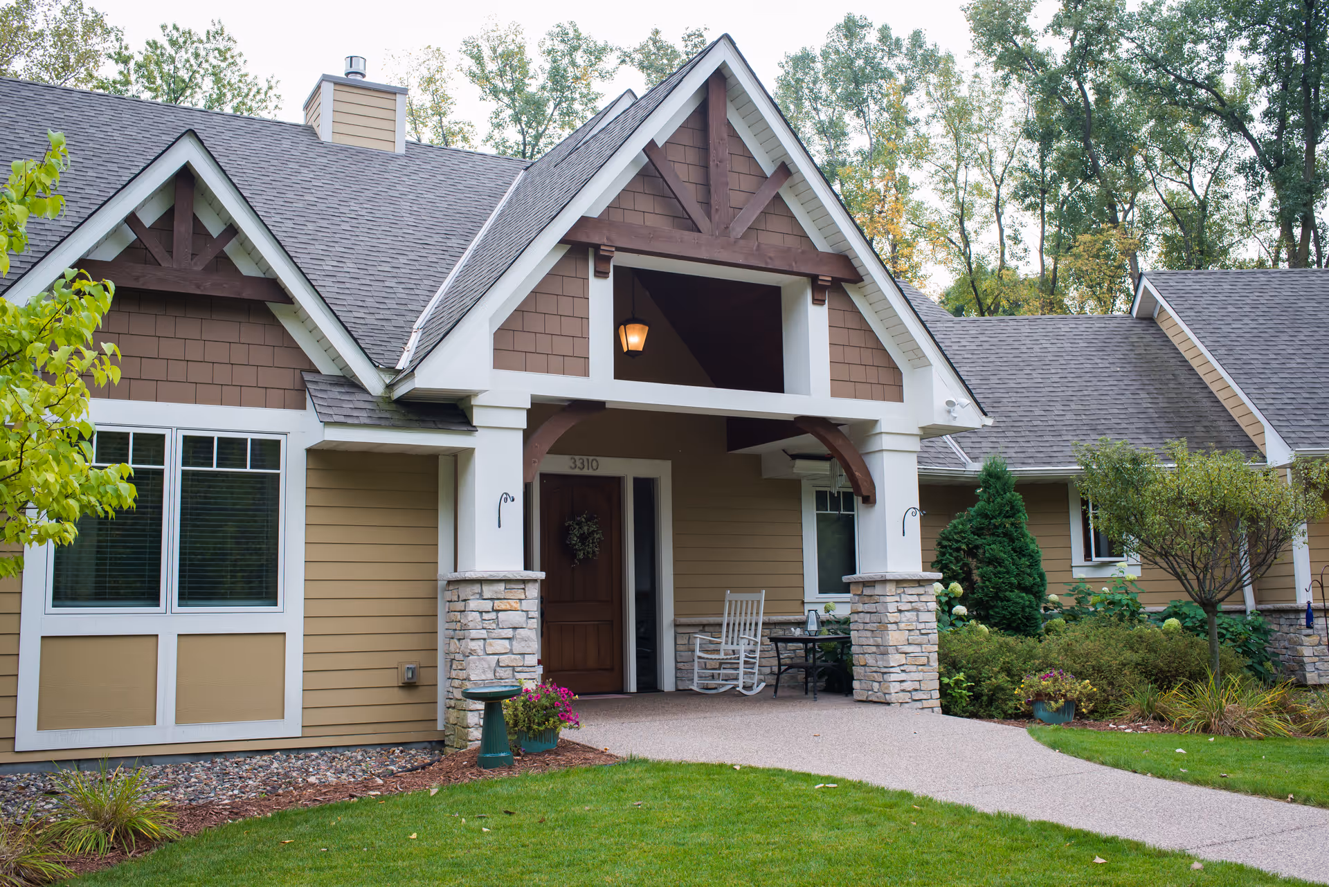 Front exterior view of a single-story senior living facility building with a covered entrance, stone pillars, a wooden door with a wreath, rocking chairs on the porch, and well-maintained landscaping including grass, shrubs, and trees.