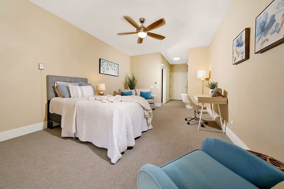 A spacious bedroom with beige walls and carpeted floor featuring a bed with white bedding and multiple pillows, a small sofa with blue cushions, a wooden desk with a chair, a floor lamp, and wall art. A ceiling fan with a light is mounted above the bed.