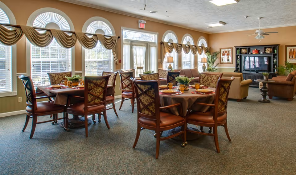 A spacious and well-lit common area in a senior living facility with round dining tables set with plates, glasses, and napkins. The room features large arched windows with decorative valances, comfortable upholstered chairs, and a seating area with sofas and armchairs near a TV cabinet. The walls are painted a warm beige color, and there are plants and framed artwork adding to the cozy atmosphere.