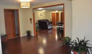 Interior view of a senior living facility showing a hallway with wooden floors and several potted plants. The hallway leads to a living room area with a dark leather sofa and a dining table with chairs. There are wooden doors and a ceiling light fixture visible.