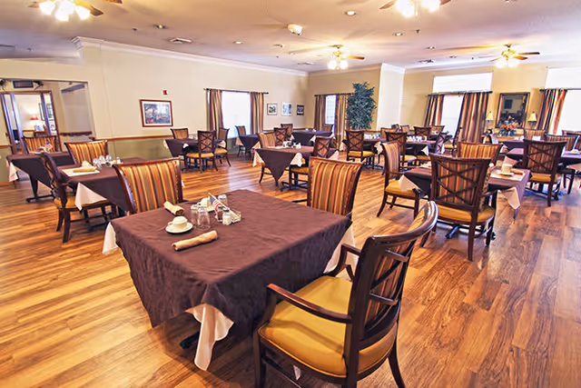 Spacious dining room with multiple tables covered in dark brown tablecloths and set with cups, napkins, and condiments. The room has wooden flooring, several windows with curtains, ceiling fans, and framed pictures on the walls.