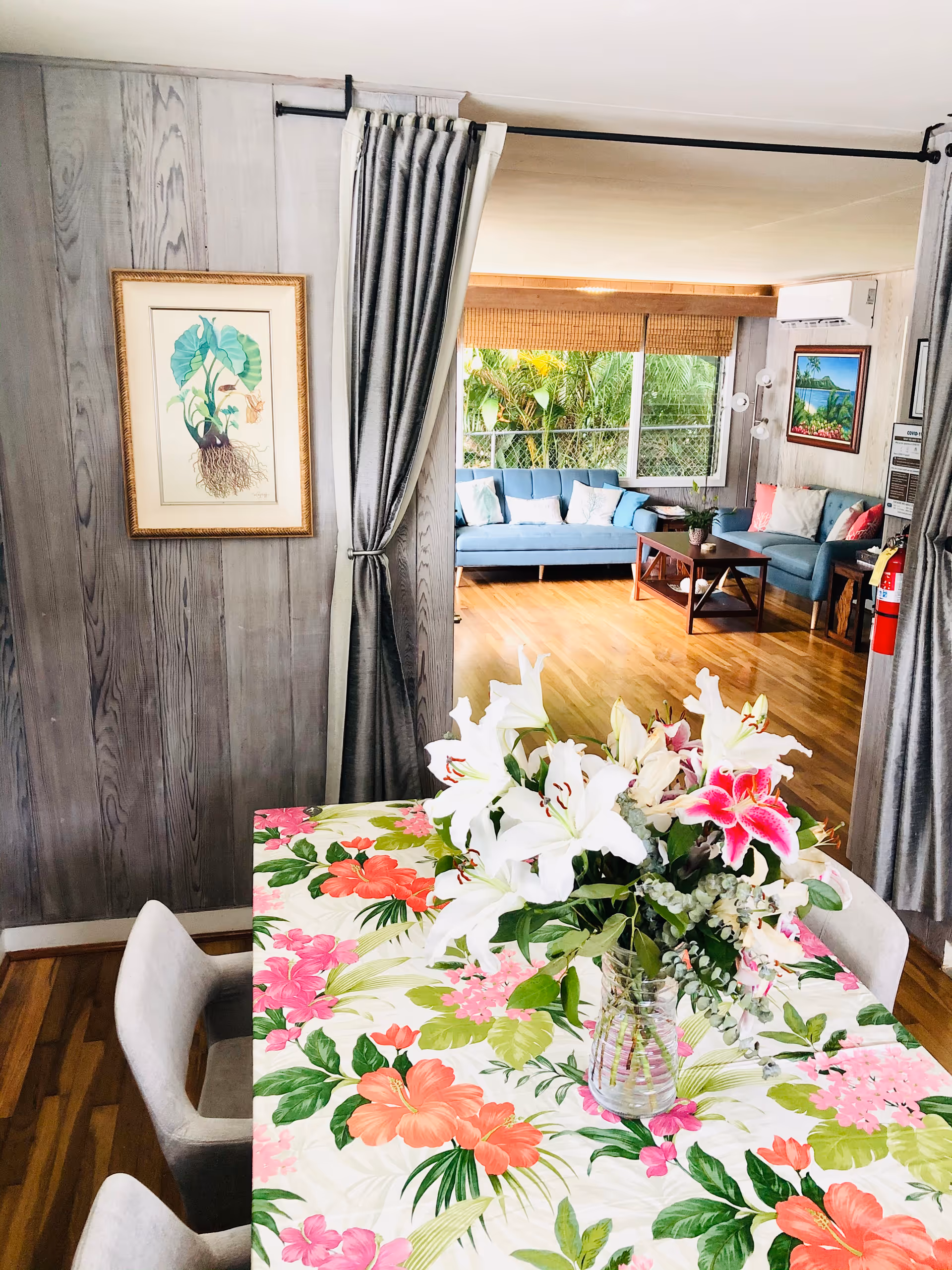 Dining table with a colorful floral tablecloth and a vase of lilies in the foreground, leading into a sunlit living room with blue sofas.