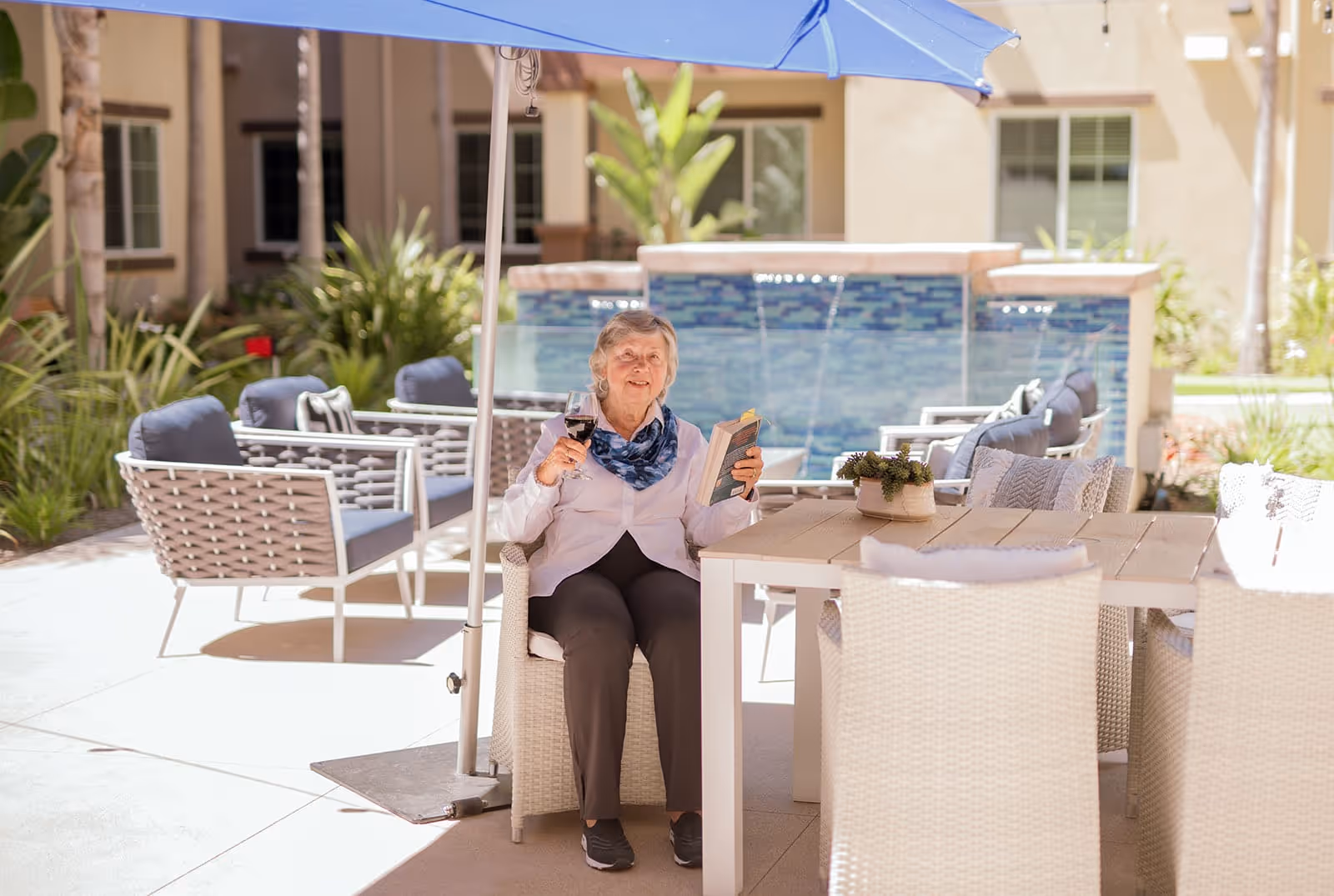 An elderly woman sitting outdoors under a blue umbrella at a patio table, holding a glass of wine in one hand and a book in the other. The patio features cushioned chairs, a water feature in the background, and surrounding greenery.