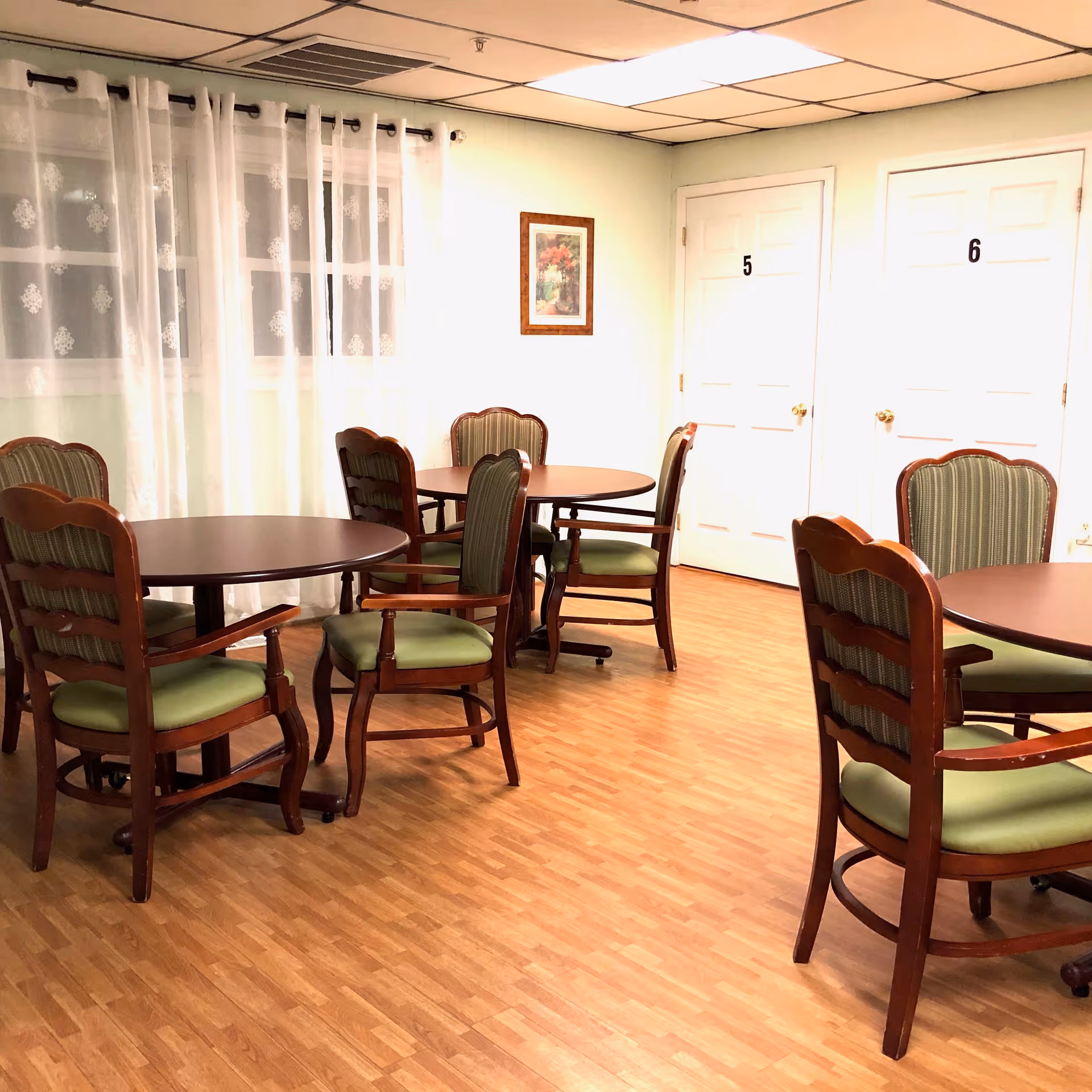 A room with wooden round tables and green cushioned chairs arranged on a wood-patterned floor. Sheer white curtains cover the windows on the left wall, and two white doors labeled with numbers 5 and 6 are visible on the right wall. A framed floral picture hangs between the doors.