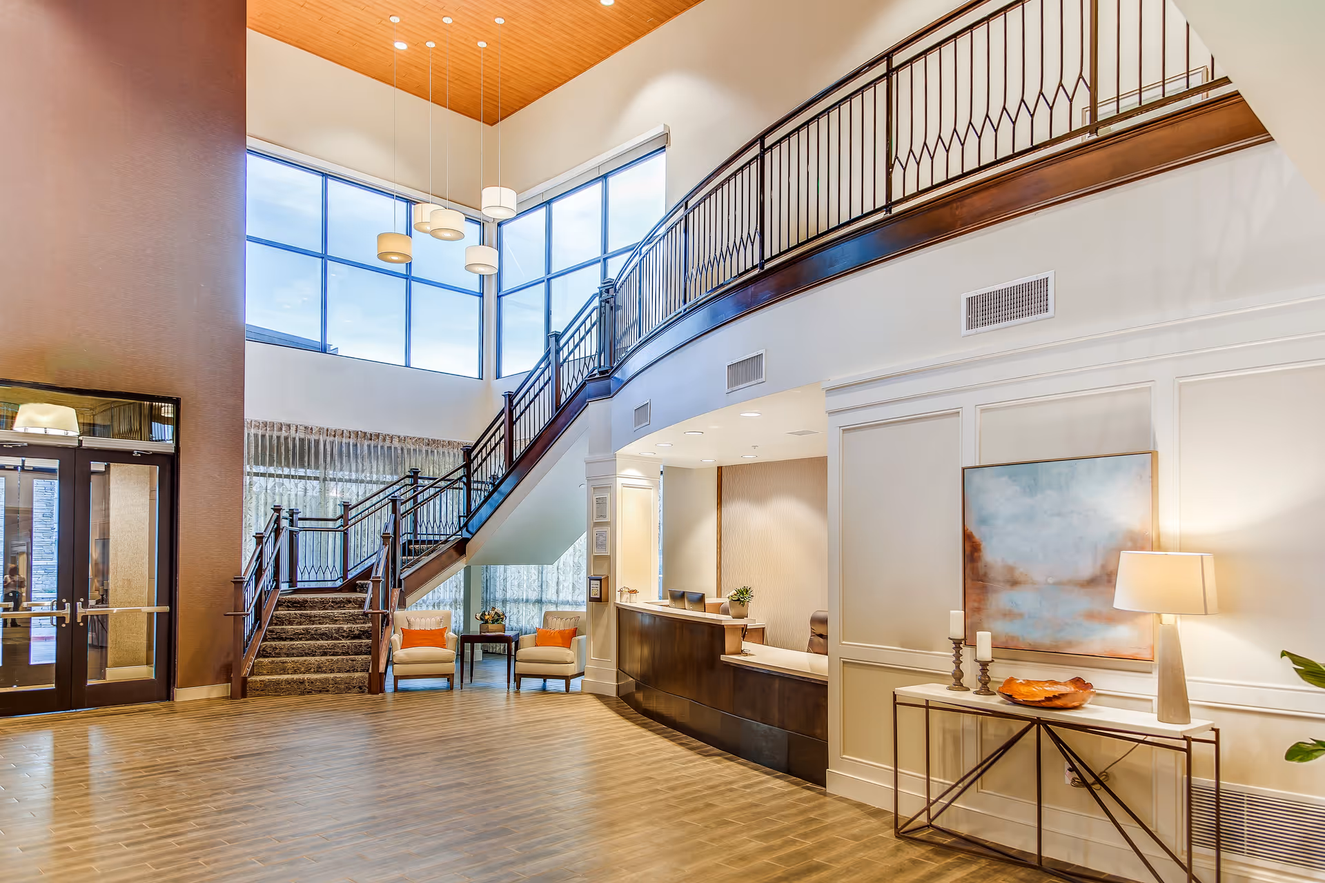 Spacious and well-lit lobby area with a wooden staircase leading to an upper floor, large windows letting in natural light, a reception desk with chairs behind it, two armchairs with orange cushions beside a small table, and a console table with a lamp, candles, and artwork on the wall.