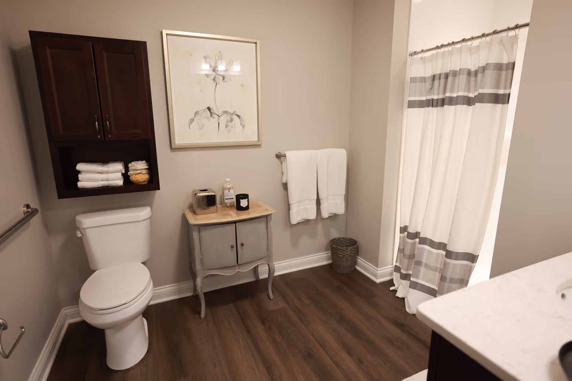 A clean and modern bathroom featuring a white toilet, a small wooden cabinet with toiletries on top, a wall-mounted cabinet with folded towels, a towel rack with two white towels, a wicker wastebasket, and a shower with a white curtain that has gray stripes. The floor is wood-style and the walls are painted beige.