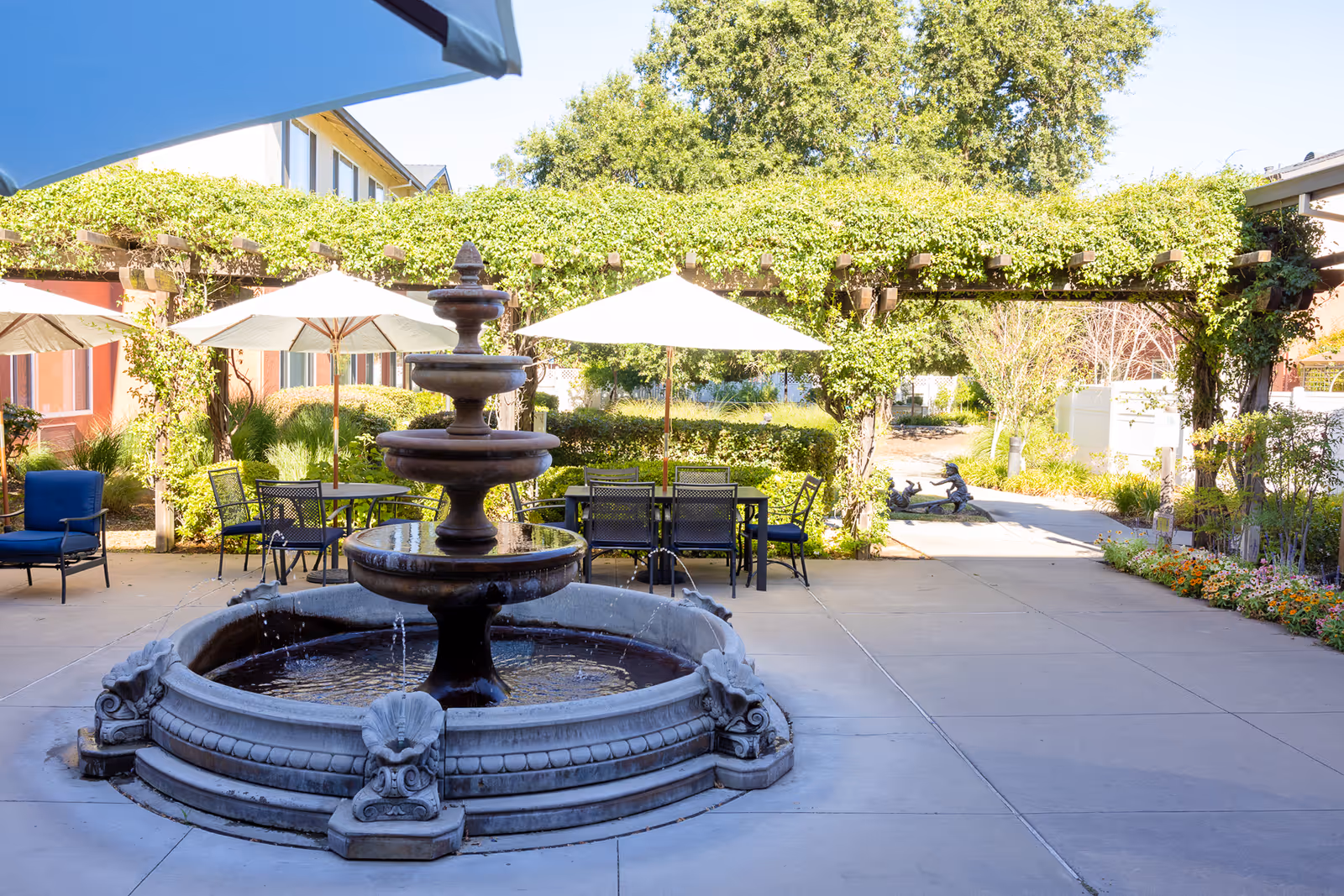 Outdoor patio area at Carlton Senior Living Sacramento featuring a large tiered stone fountain in the foreground, several tables with umbrellas, chairs, and a pergola covered with green vines. There are trees and plants surrounding the paved area under a clear blue sky.