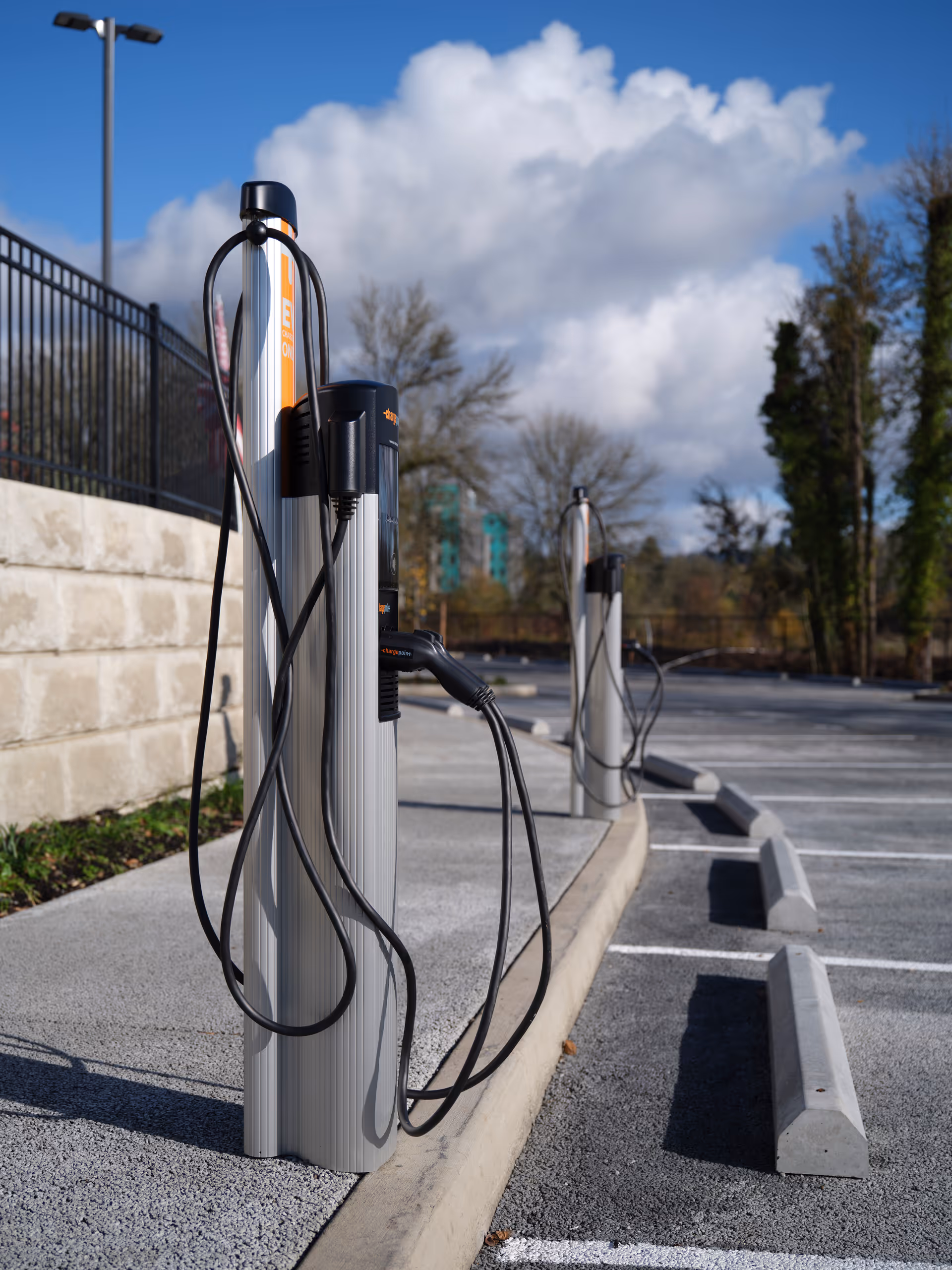 Electric vehicle charging stations in an empty parking lot on a sunny day with a partly cloudy sky and trees in the background.