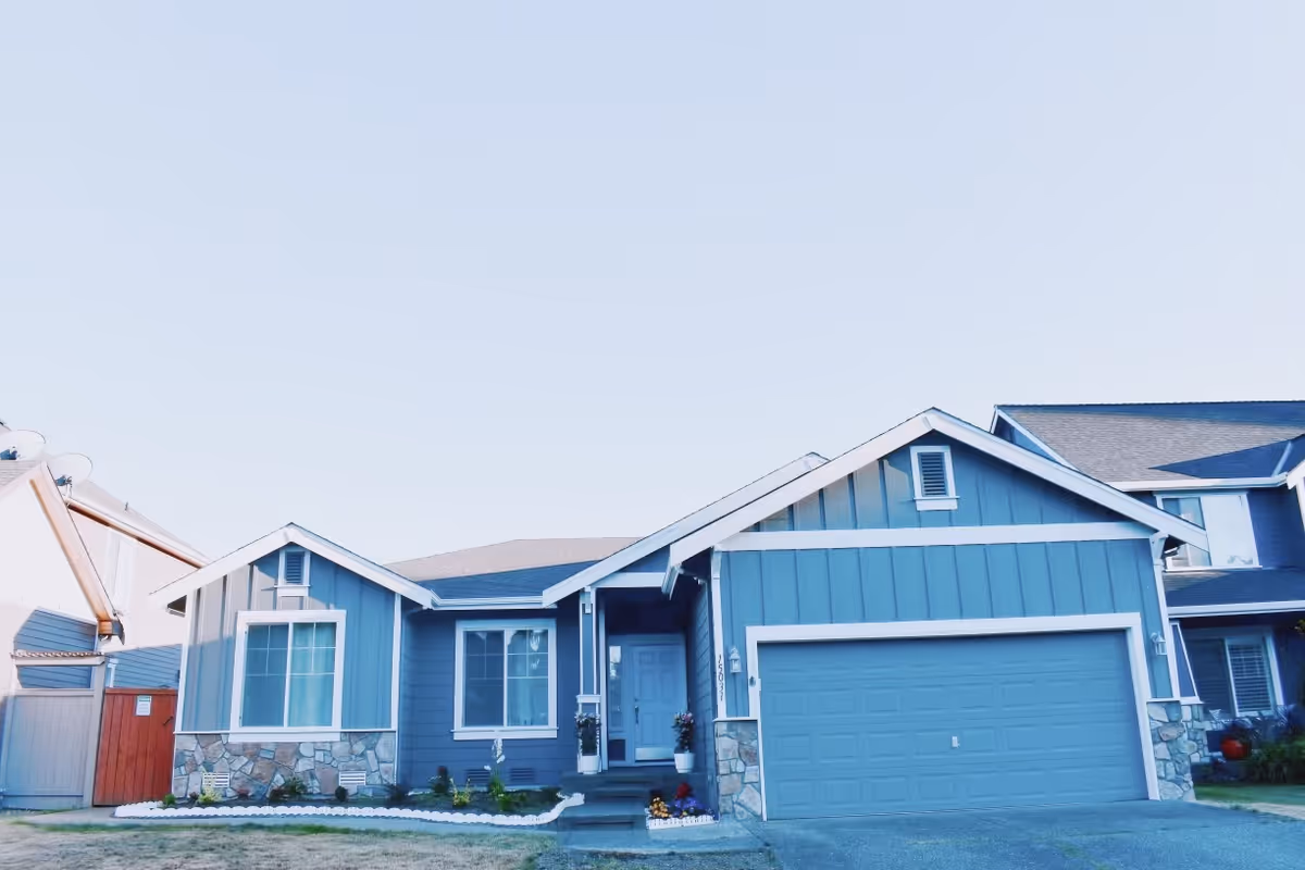 Front exterior view of a single-story blue house with white trim, a two-car garage, and a small garden area with flowers near the entrance.
