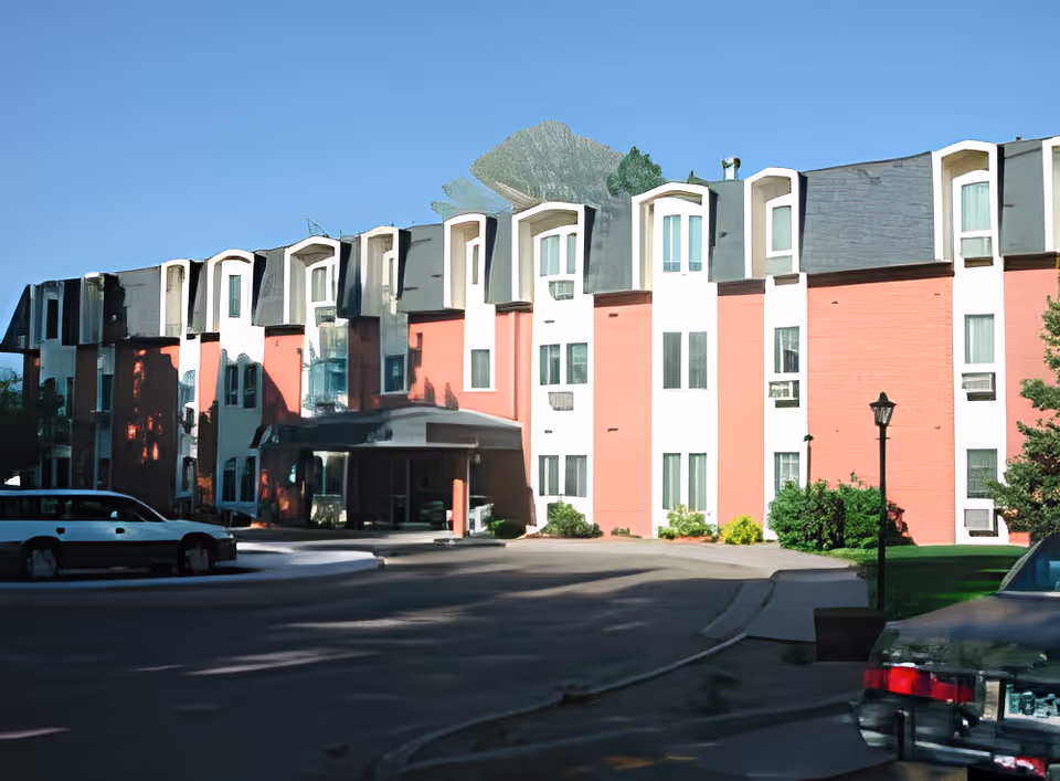 Exterior view of a multi-story senior living facility building with a red and white facade, multiple windows, and a covered entrance. There are cars parked in front and a clear blue sky above.