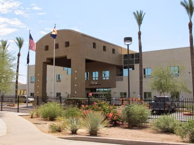 Exterior view of the Arizona State Veteran Home in Phoenix, showing a two-story building with a covered entrance, several windows, three flagpoles with flags, desert landscaping with bushes and palm trees, and a black metal fence in front.