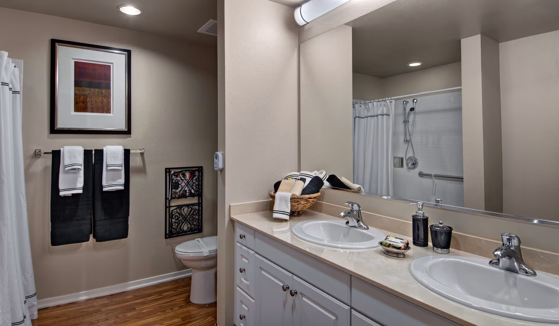 A clean and modern bathroom featuring a double sink vanity with a large mirror above it. The countertop holds a basket with rolled towels and soap dispensers. In the background, there is a toilet with a decorative wall hanging above it, two black towels with white hand towels hanging on a towel rack, and a shower area with a white curtain and a handheld showerhead.