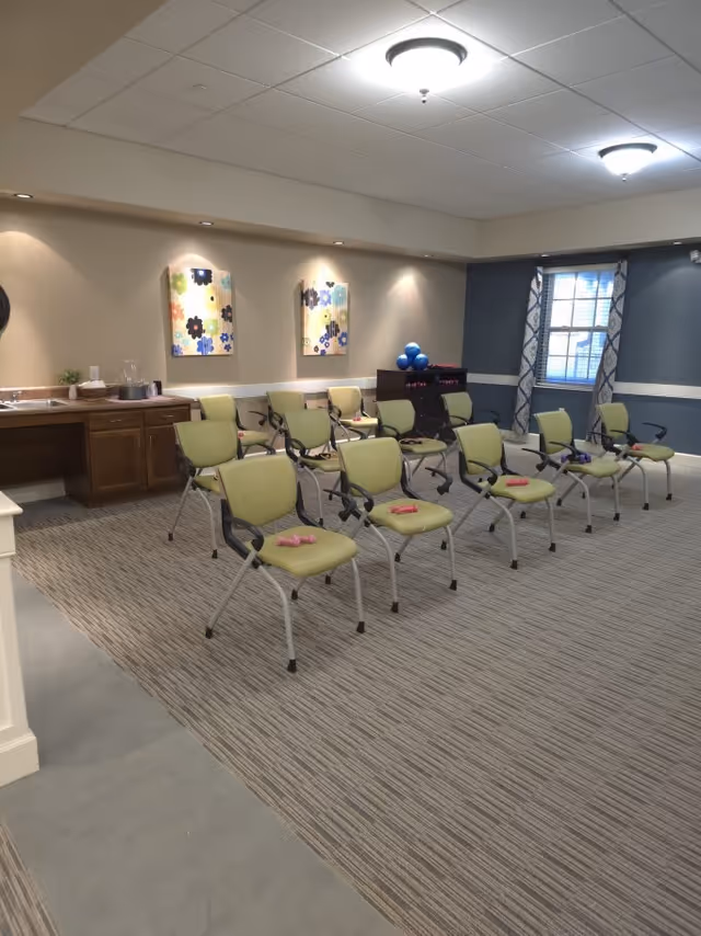 A room with multiple green chairs arranged in rows, each chair has a small pink dumbbell on the seat. The room has a carpeted floor, beige and blue walls, two colorful floral paintings on the wall, a window with patterned curtains, and a cabinet with blue exercise balls on top.