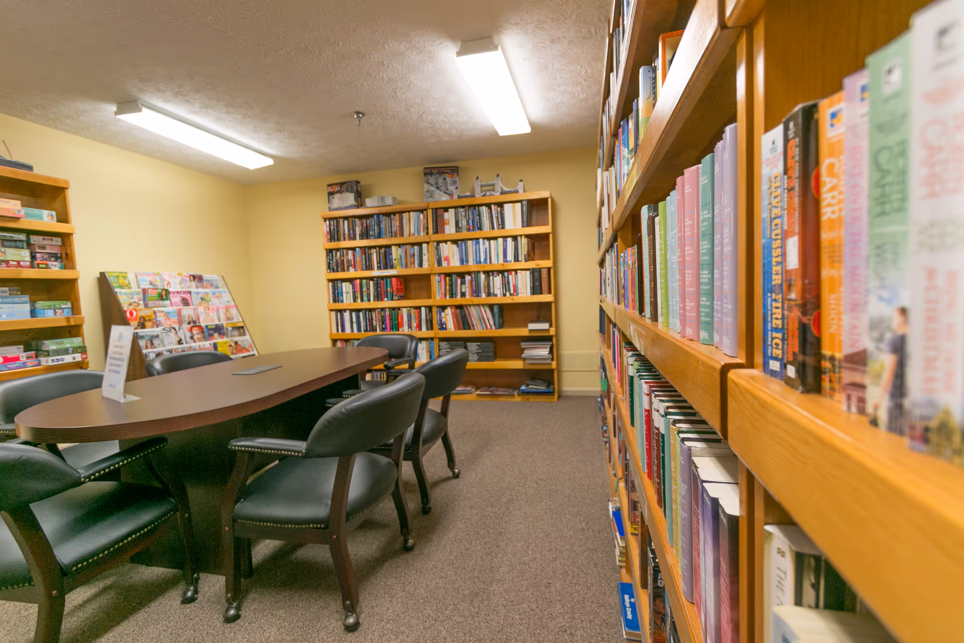 A cozy interior library/reading room with wooden bookshelves, an oval table surrounded by chairs, and a magazine rack.